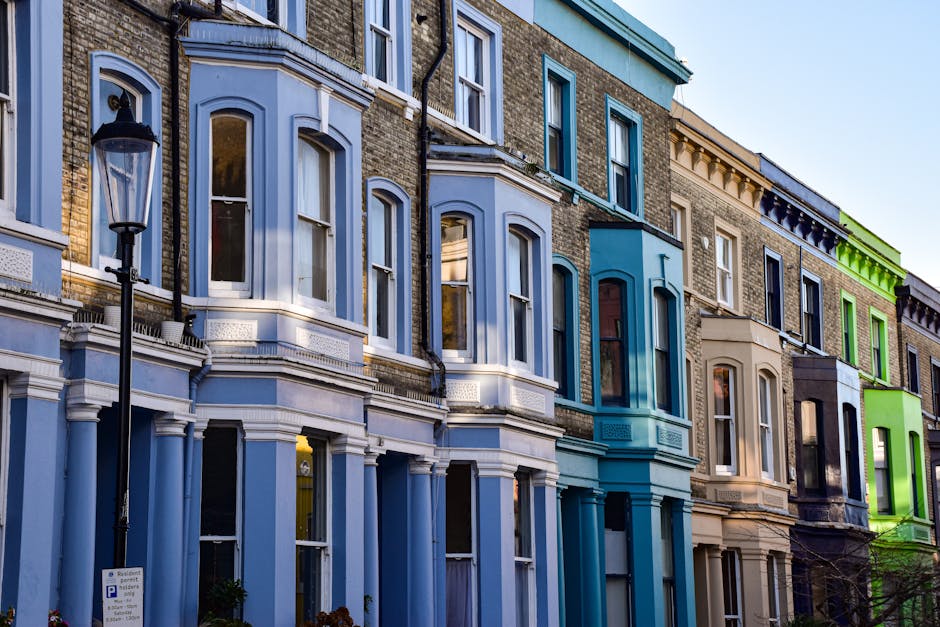 A row of multi-storey terraced houses with Victorian-style architecture, featuring bay windows and decorative mouldings, painted in various shades including white, blue, beige, and green. The buildings are situated along a street, with a black lamppost visible on the pavement in front. The windows are large, with some reflecting the sky and surroundings. The scene is captured during daylight, highlighting the different colours and architectural details of the residences. Inside one of the houses, visible through the open door, are cardboard boxes and packing materials, indicating a home relocation or moving process. The loading area shows a move being prepared, with a moving truck likely nearby, as part of a furniture transport or packing and moving service provided by Finsbury Park Movers.