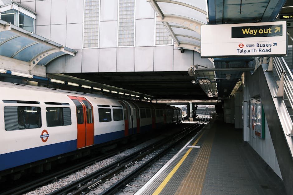 Inside a London underground station, a train with white, orange, and blue exterior panels is stationary on the tracks, illuminated by overhead lighting. The platform features a textured yellow safety strip along the edge and is partially shaded by a modern, curved metal and glass canopy overhead. To the right, there is an outdoor staircase leading to an upper level, with signage pointing towards bus services and Talgart Road, indicating the station's exits and transport connections. The station environment appears clean and organized, with a focus on safe, efficient passenger movement. This setting is relevant to house removals and moving logistics, illustrating the transportation infrastructure that supports relocating with services like those offered by Finsbury Park Movers. The scene captures the typical environment where household items may be transported via public transit before or after a home relocation, emphasizing the importance of coordination in moving processes.
