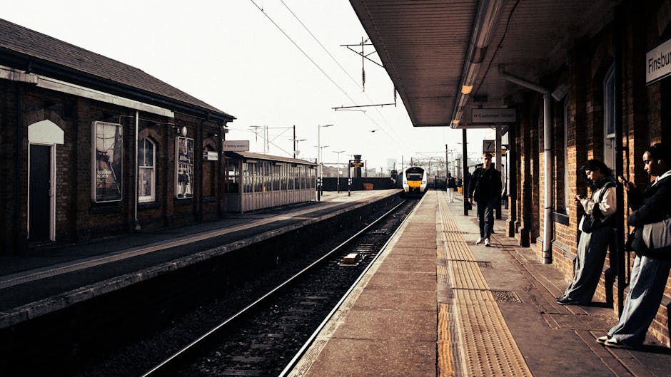 At a UK train station platform near Finsbury Park, several individuals are waiting alongside a brick-building structure featuring large windows and a covered shelter area. The platform surface includes tactile paving strips for visually impaired passengers. Close to the edge of the platform, a couple of people are standing, some engaged with their mobile devices, while others are observing the approaching train. A yellow and white train is visible in the background, arriving on the tracks, which are equipped with overhead electric lines and support poles. On the opposite side of the tracks, another platform is visible with additional waiting passengers and station signage. The scene is lit with natural daylight, suggesting daytime conditions. As part of a home relocation or moving process, Finsbury Park Movers could be involved in coordinating transportation, including the safe loading and unloading of household items onto the train or vehicles, ensuring efficient logistics in moving furniture and packed boxes during the packing and moving phase near Finsbury Park Station.