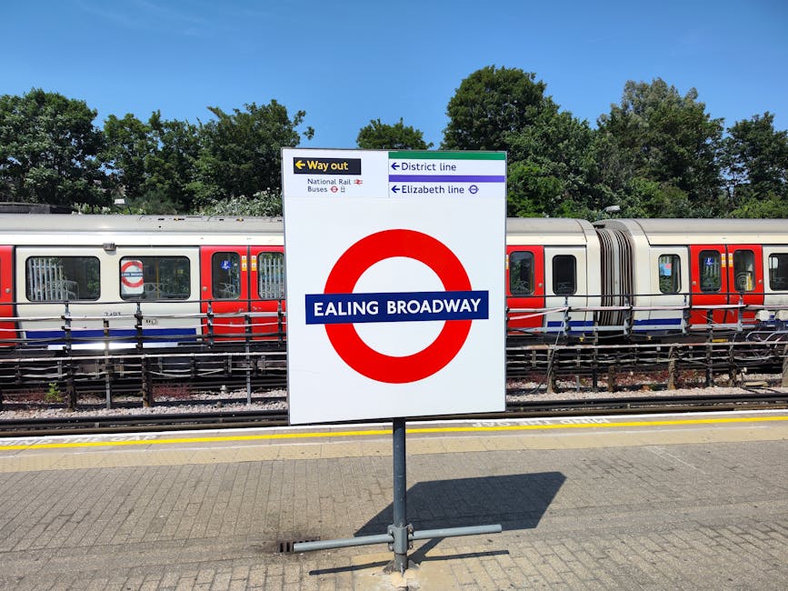 Inside a London underground station, a train with white, orange, and blue exterior panels is stationary on the tracks, illuminated by overhead lighting. The platform features a textured yellow safety strip along the edge and is partially shaded by a modern, curved metal and glass canopy overhead. To the right, there is an outdoor staircase leading to an upper level, with signage pointing towards bus services and Talgart Road, indicating the station's exits and transport connections. The station environment appears clean and organized, with a focus on safe, efficient passenger movement. This setting is relevant to house removals and moving logistics, illustrating the transportation infrastructure that supports relocating with services like those offered by Finsbury Park Movers. The scene captures the typical environment where household items may be transported via public transit before or after a home relocation, emphasizing the importance of coordination in moving processes.