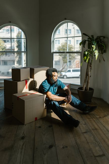 A man with dark skin and short black hair, dressed in a maroon t-shirt and bright orange trousers, stands inside a room surrounded by numerous cardboard moving boxes of various sizes, some sealed with