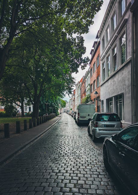A narrow residential street with a cobblestone surface, lined with multi-storey buildings featuring large windows, some with white frames and others with grey or brick facades. Several parked vehicles