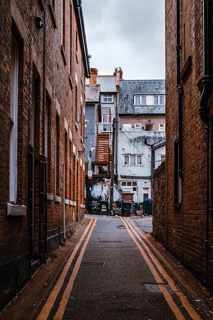 A narrow residential street with a cobblestone surface, lined with multi-storey buildings featuring large windows, some with white frames and others with grey or brick facades. Several parked vehicles