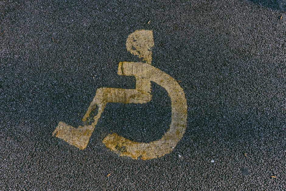 A close-up view of a wheelchair accessibility symbol painted on a dark asphalt surface, showing a stylized figure seated in a wheelchair with a rounded wheel below. The paint is weathered and slightly faded, with some areas chipped. The surrounding asphalt has small stones and debris scattered across it, indicating outdoor street or pavement area. This image captures the clear marking used for accessible parking zones, relevant to house removals and parking considerations during furniture transport and home relocation processes, as discussed in Finsbury Park Movers' page about Seven Sisters Road removals access and parking tips.