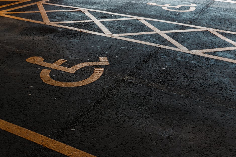 Close-up view of a designated accessible parking space on an asphalt surface, featuring a large, orange chalk outline of a wheelchair symbol in the foreground. Surrounding the parking bay are different painted lines and markings indicating parking boundaries, with some of the lines extending into an adjacent area designated for vehicle or equipment loading during home relocation or furniture transport. The parking space appears empty and is located near a building entrance or loading zone, which is typical for moving services such as those provided by Finsbury Park Movers, that assist with packing and loading objects for removals. The surface is slightly textured and shows signs of wear, with minimal debris present, and the lighting suggests daytime conditions conducive to safe moving operations near Seven Sisters Road, FINSBURY PARK.